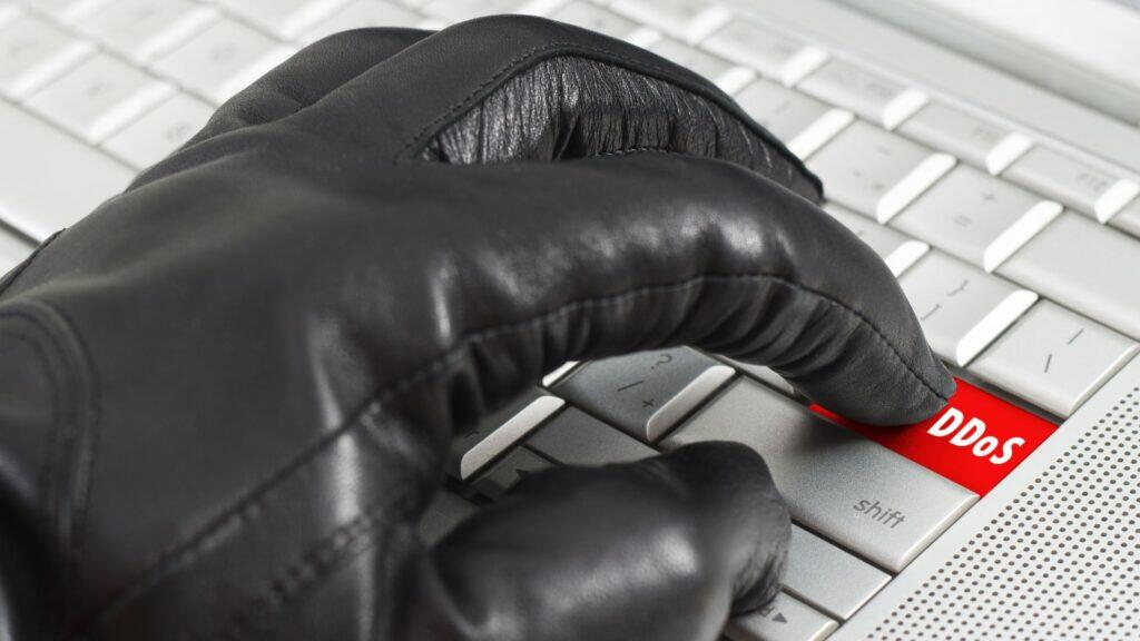 Eine Person in einem Lederhandschuh drückt einen Knopf auf einem Laptop. Symbolbild für DDoS.